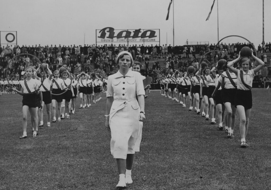 
Ćwiczenia dziewcząt z obręczami i piłką na murawie Stadionu Wojska Polskiego. Na koronie widoczna reklama fabryki obuwia "Bata".